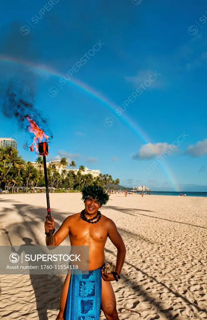 Hawaii, Oahu, Waikiki Beach, A Local Hawaiian Man Wears A Traditional Outfit, Holds A Torch With Rainbow In Background.