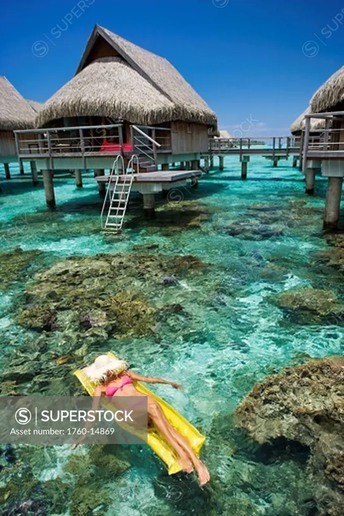 French Polynesia, Moorea, Woman sunbathing on inflatable raft over ocean reef, Luxury resort bungalows over ocean.