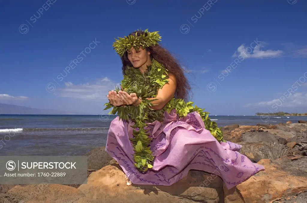 Hula dancer with haku lei in traditional outfit on shoreline, ocean background