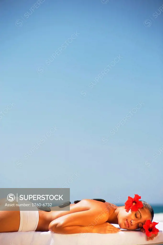 Hawaii, Topless girl laying down next to ocean on massage table with stones on her back, red hibiscus behind her ear 