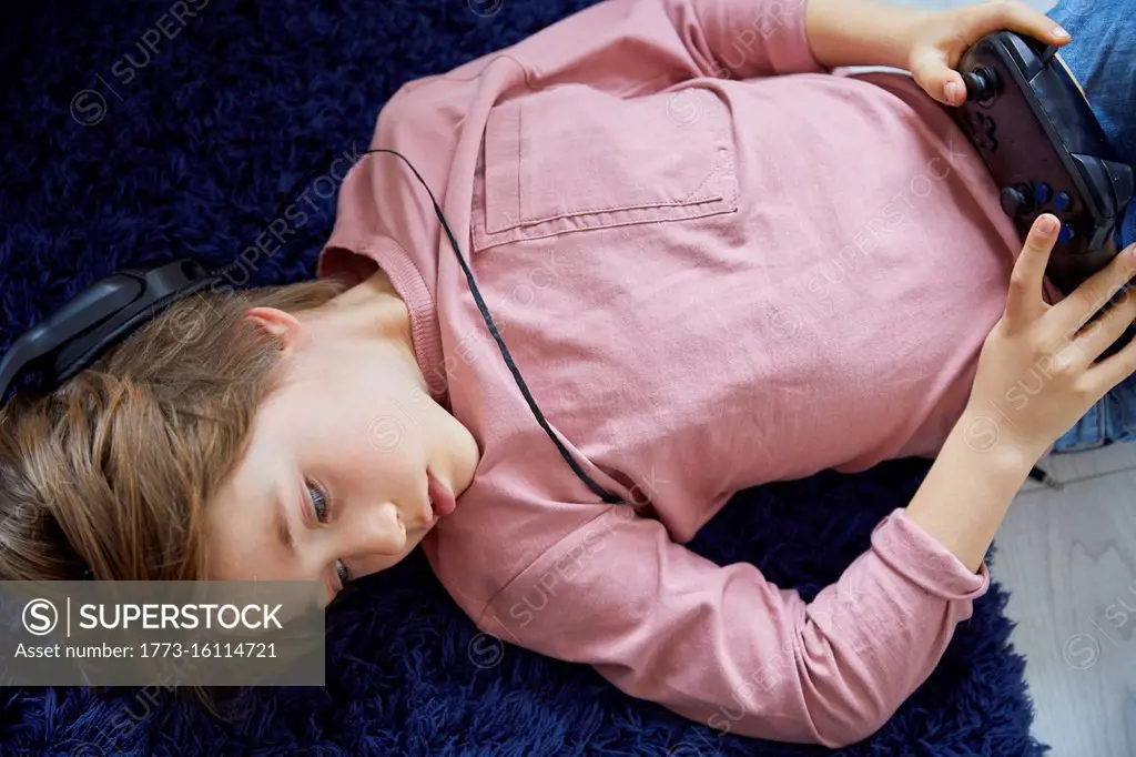 Boy wearing pink T-Shirt lying on his back on blue fluffy rug, holding game console controller.