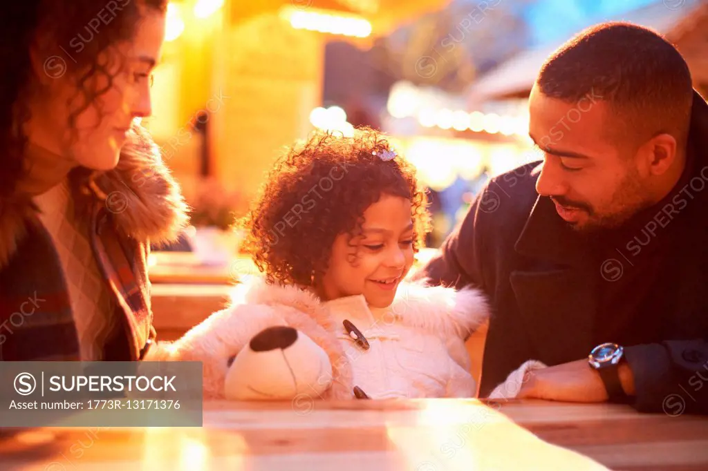 Girl at amusement park holding teddy bear sitting at table with parents