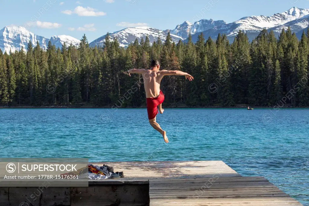 A man in a red bathing suit jumps off a dock into the clear waters of Annette Lake with the snowy Canadian Rocky Mountain in the distance in Jasper Na...