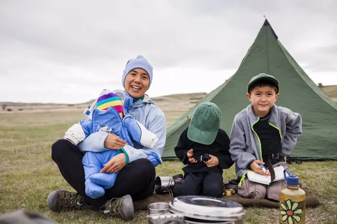 A family consisting of a Japanese American woman holding her 6 month baby girl, and 4 year old and 6 year old Japanese American boys sit in front of their tent while camping in a primitive campsite, Sage Creek Campground, Badlands National Park, South Dakota.