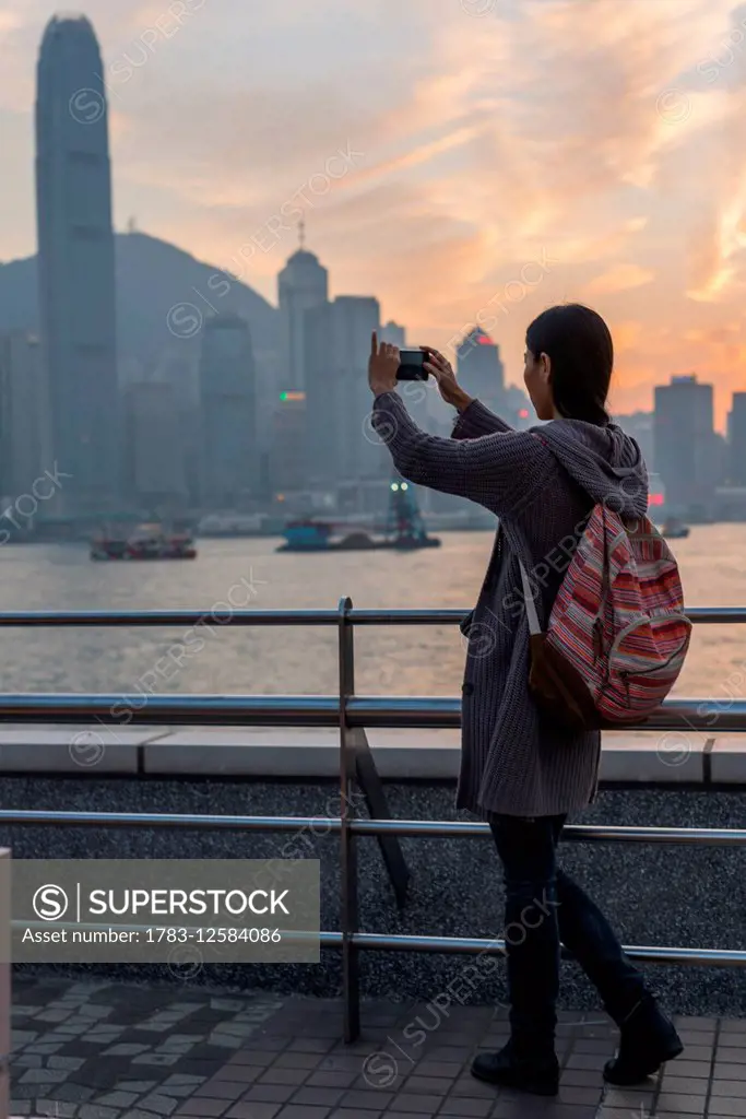 A young woman takes a picture with her camera of the harbour and Hong Kong skyline at sunset, Kowloon; Hong Kong, China