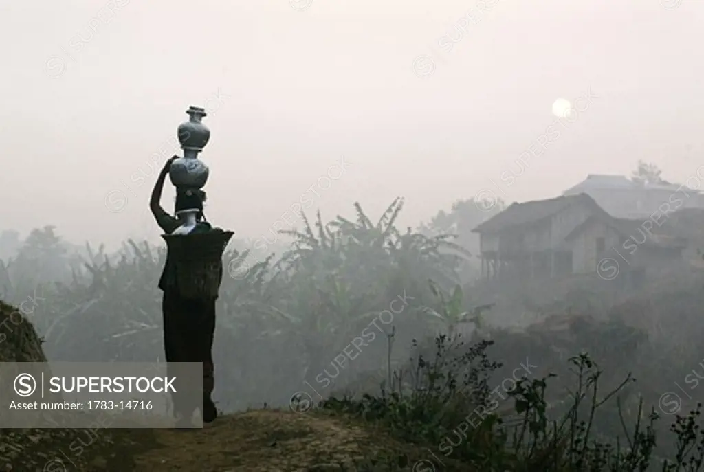 Bru tribeswoman carrying water pots on her head at sunrise, Tripura , North East States , India . The Bru Tribe is also known as the Reang tribe.