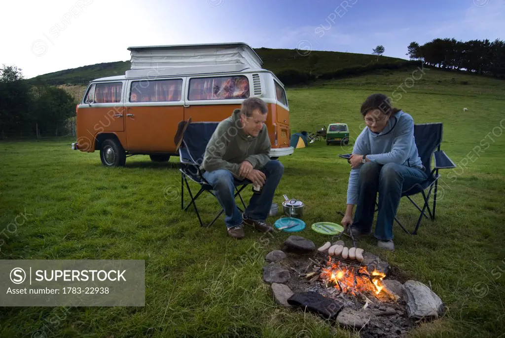 Couple cooking sausages on open fire in front of old camper van, Exmoor, England