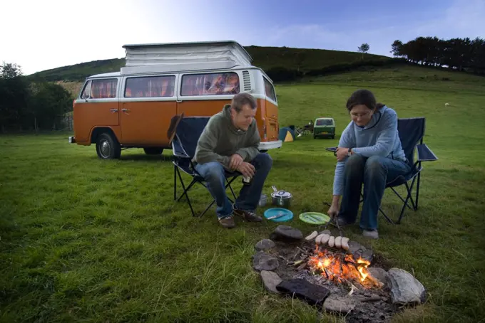 Couple cooking sausages on open fire in front of old camper van, Exmoor, England