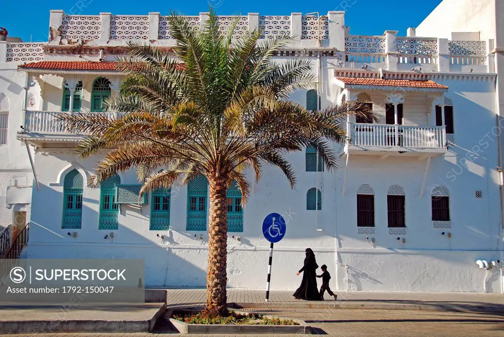 Sultanate of Oman, Muscat, Omani woman fully covered with the traditional Arabic black abbeya, walking alongside a traditional Omani house, with a pal...