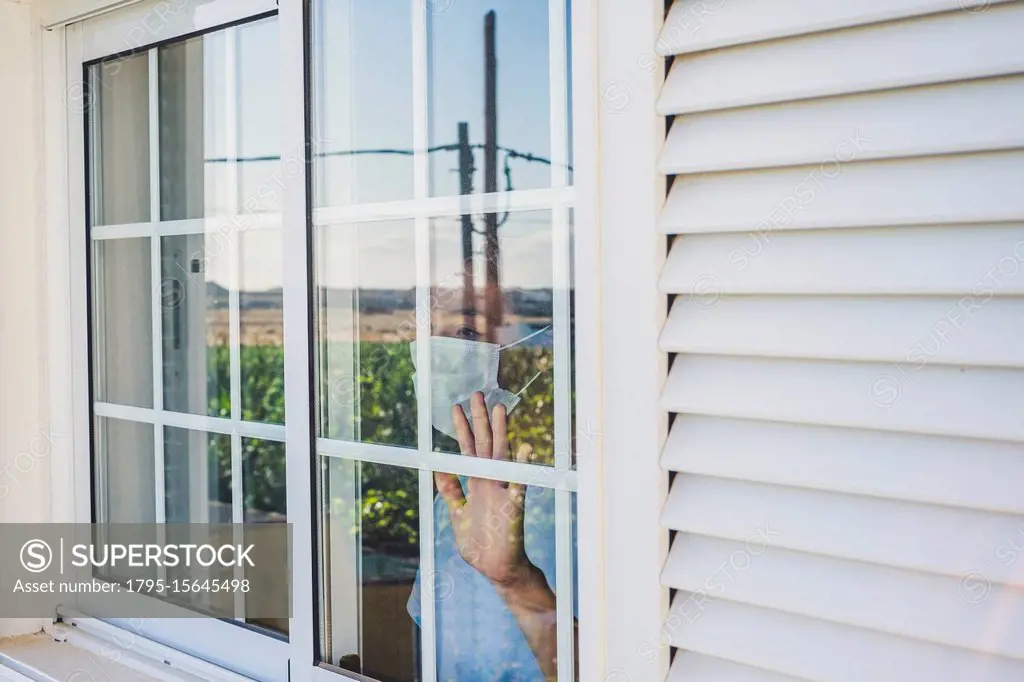 Teenage boy (16-17) wearing protective mask looking through window