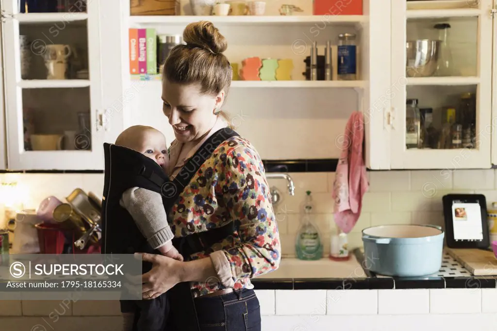 Mid adult mother with baby son in sling in kitchen