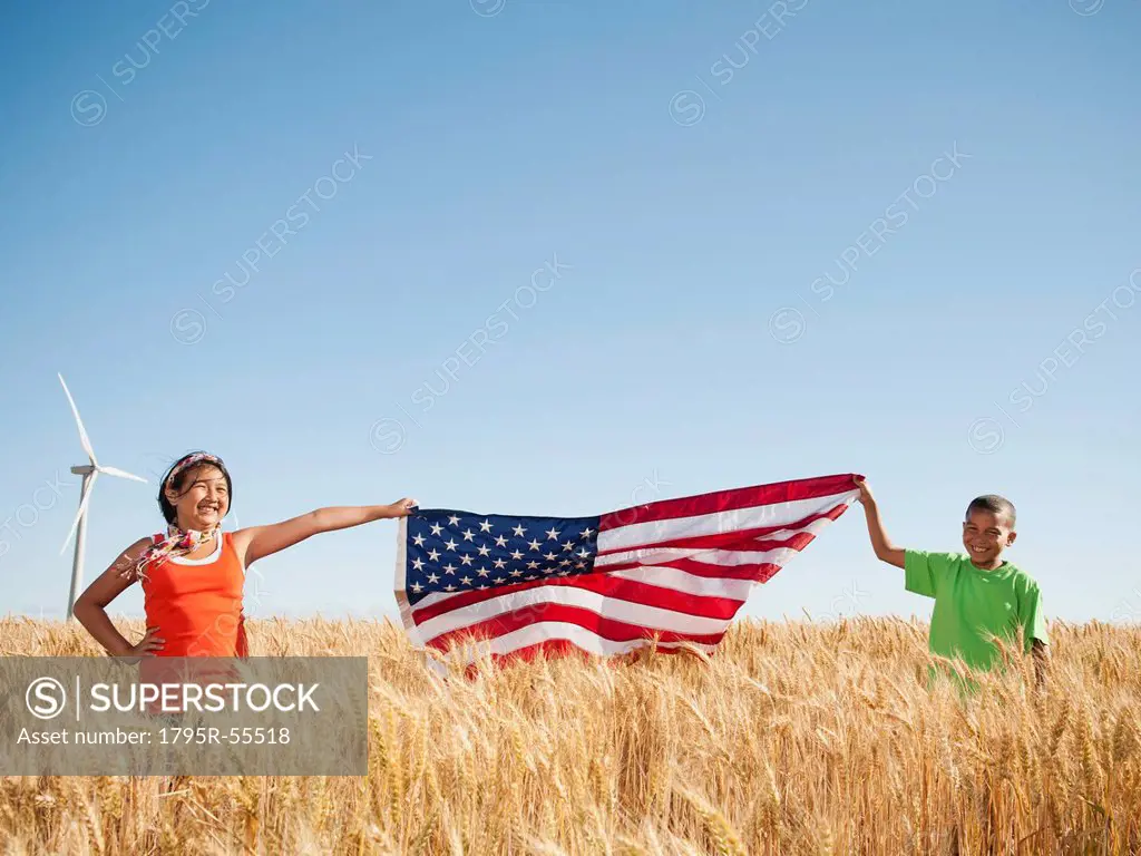 USA, Oregon, Wasco, Girl 10_110 and boy 8_9 holding american flag in wheat field, wind turbine in background