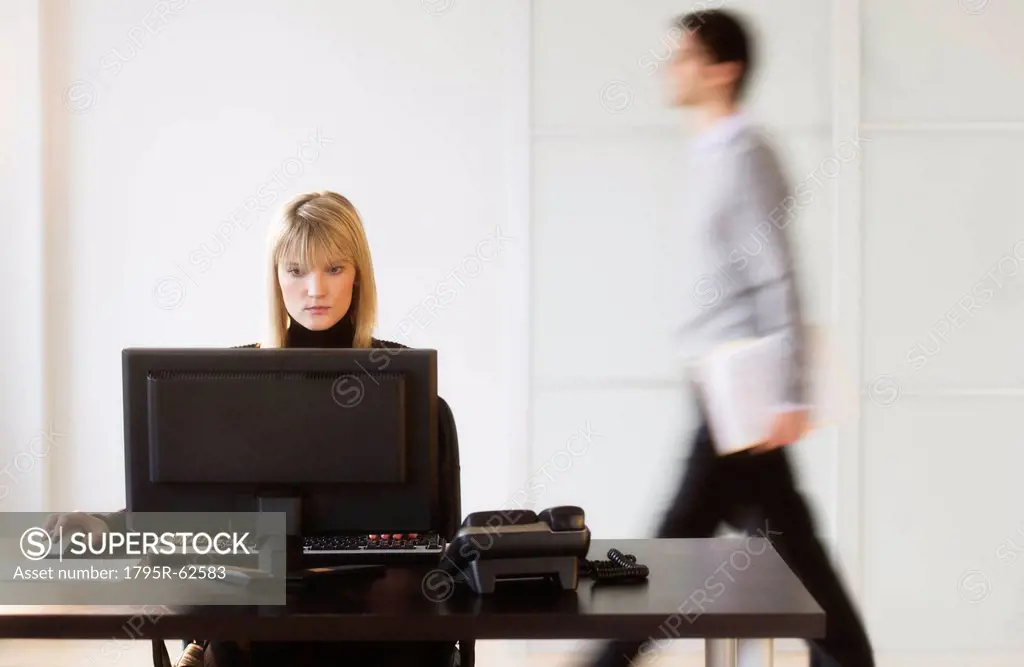 Businesswoman working on computer, businessman walking in background