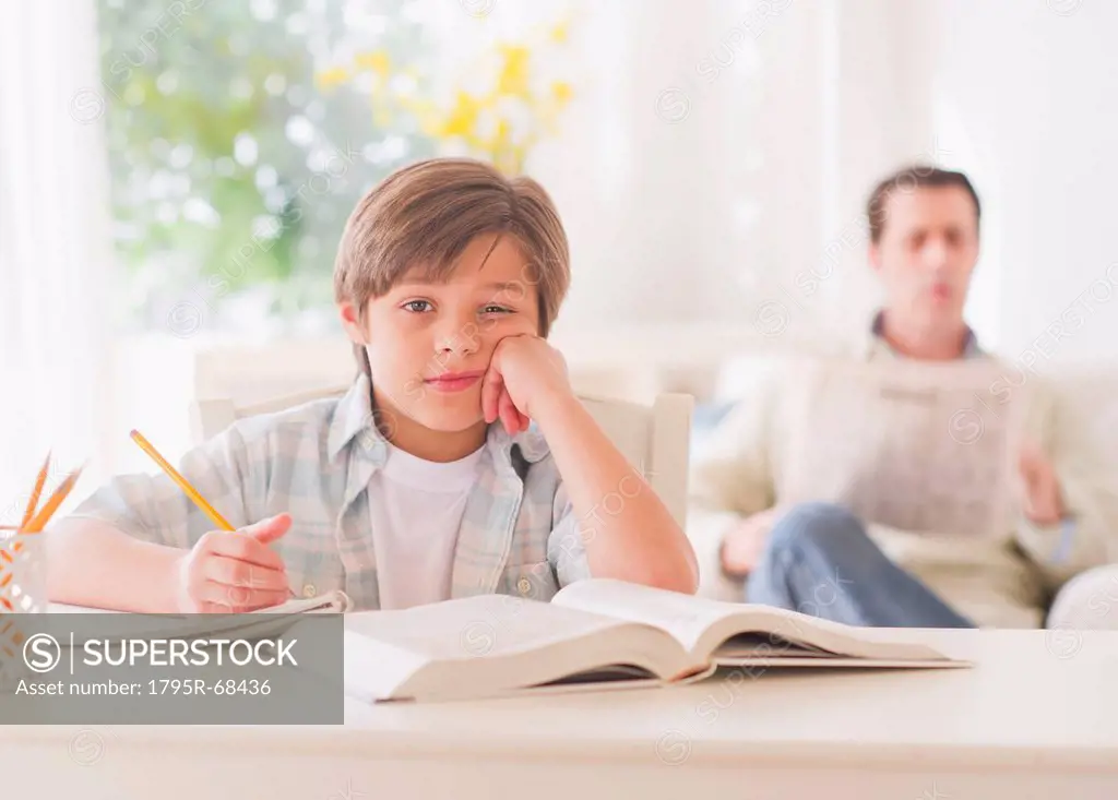 Boy 10_11 years doing homework at table with father sitting in background