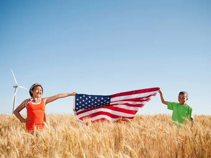 USA, Oregon, Wasco, Girl 10_110 and boy 8_9 holding american flag in wheat field, wind turbine in background