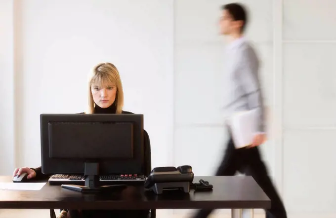 Businesswoman working on computer, businessman walking in background