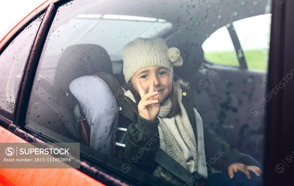 Portrait of happy little girl with wool cap doing victory sign while travelling in car