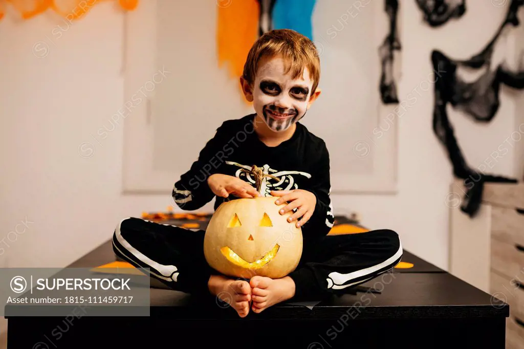 Portrait of smiling little boy with painted face and fancy dress sitting on table with Jack O'Lantern