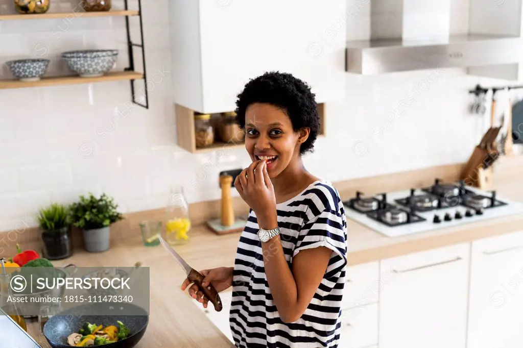 Portrait of smiling young woman cooking in kitchen at home tasting vegetables