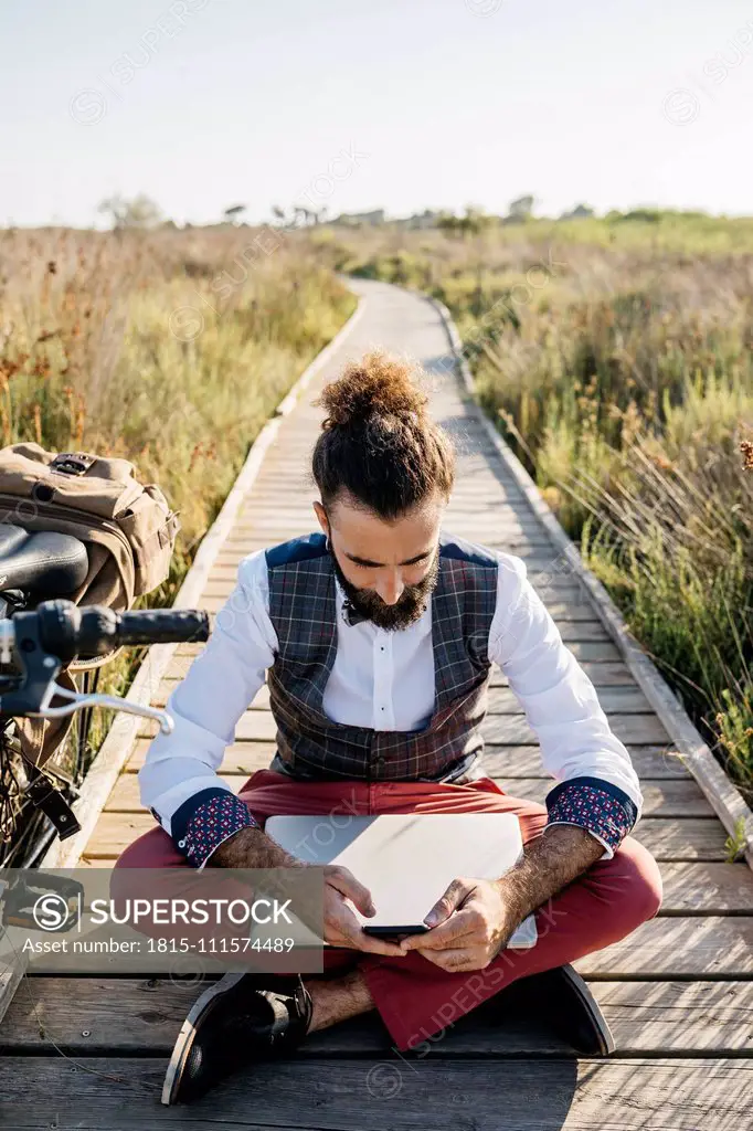 Well dressed man sitting on a wooden walkway in the countryside next to a bike with cell phone and laptop