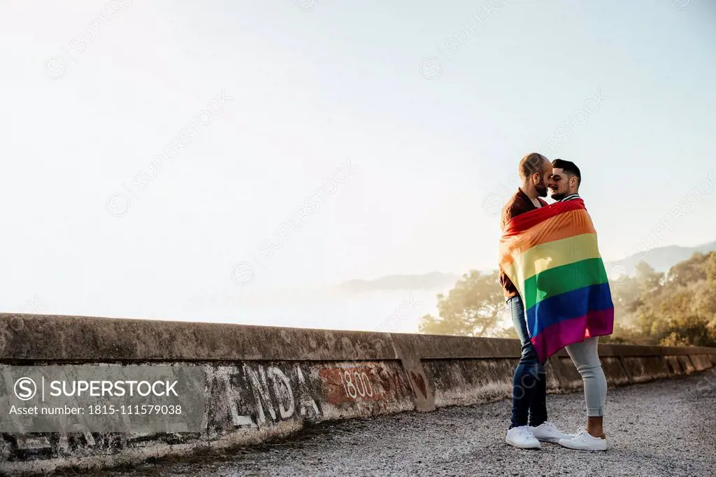 Gay couple wrapped in a gay pride flag kissing on a road in the mountains