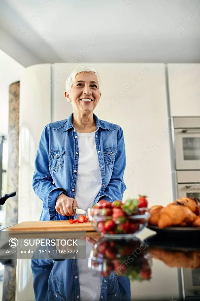 Senior woman standing in kitchen, chopping strawberries