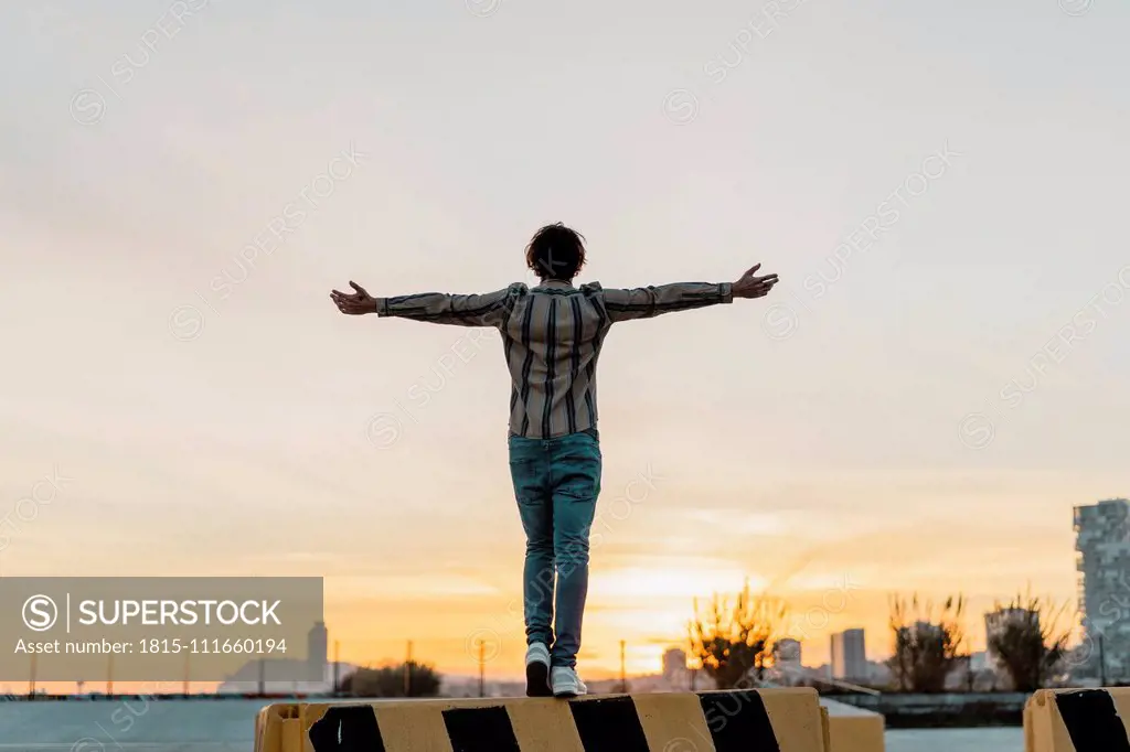 Back view of man with arms outstretched standing on barrier enjoying sunset, Barcelona, Spain