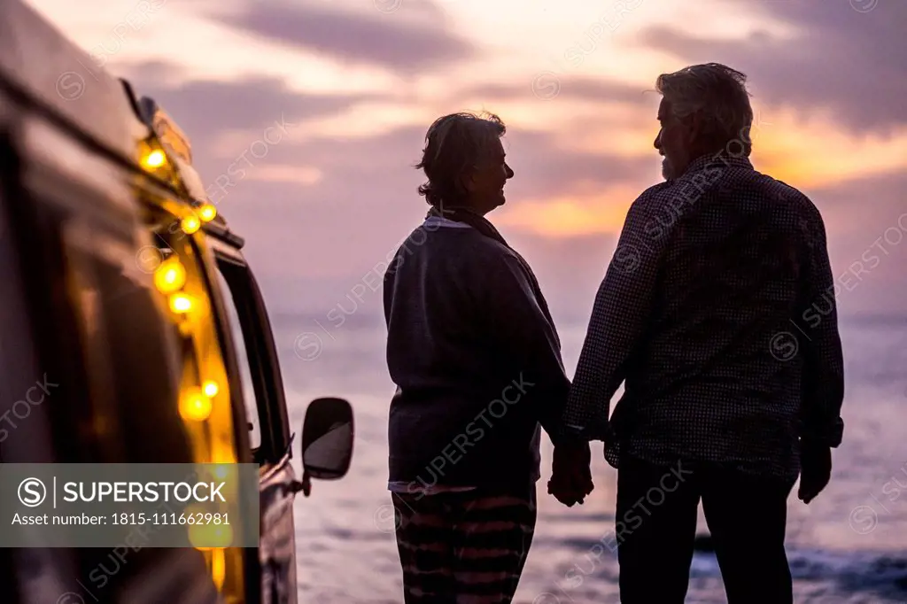 Senior couple traveling in a vintage van, watching sunset at the sea