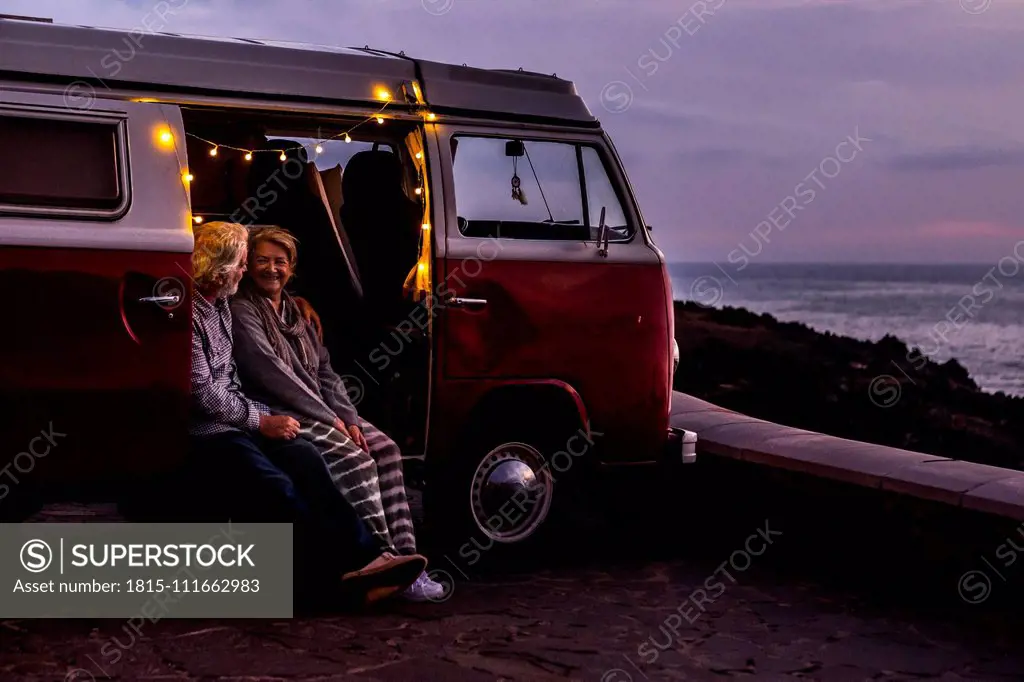 Senior couple traveling in a vintage van, watching sunset at the sea