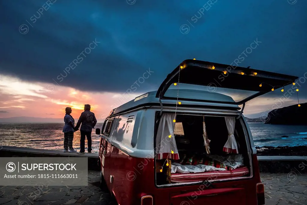 Senior couple traveling in a vintage van, watching sunset at the sea