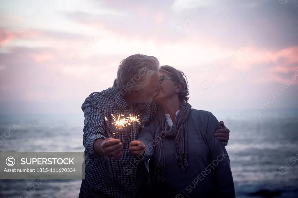 Kissing senior couple standing in front of the sea by sunset holding sparklers