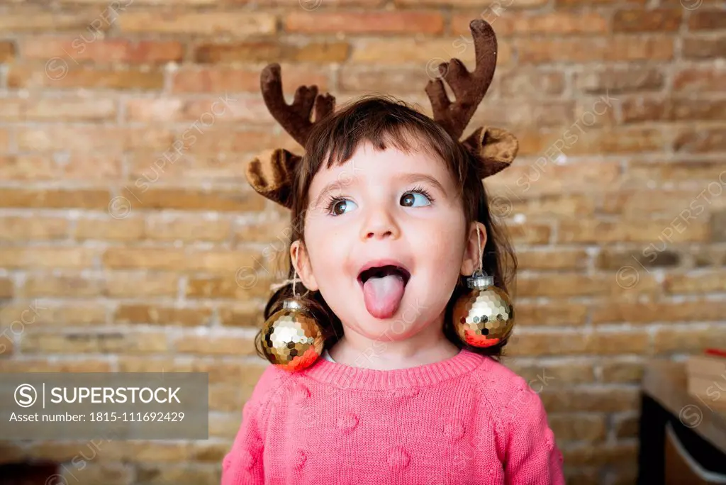 Portrait of toddler girl sticking out tongue wearing reindeer antlers headband and Christmas baubles