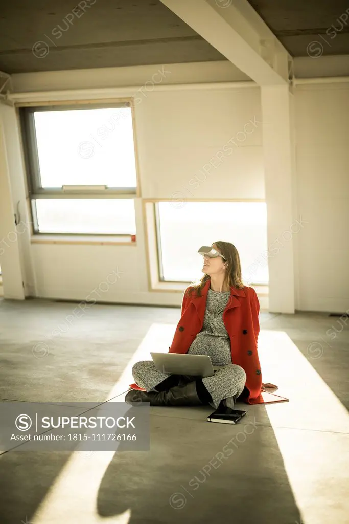 Pregnant busnesswoman sitting on floor of new office rooms, using VR goggles and laptop