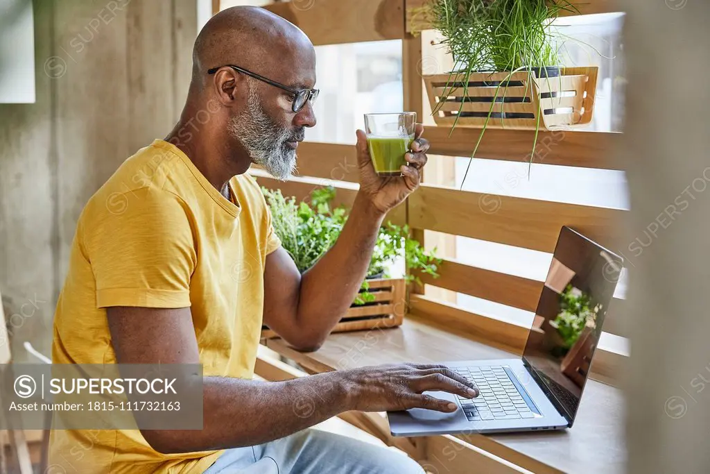 Mature businessman with smoothie using laptop at the window in office