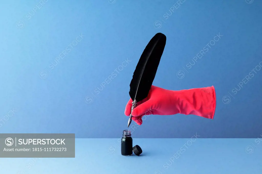 Quill pen held in hand and dipping in inkwell over blue background