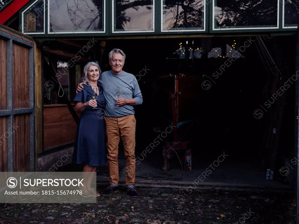 Senior couple standing in front of boathouse with glass of champagne