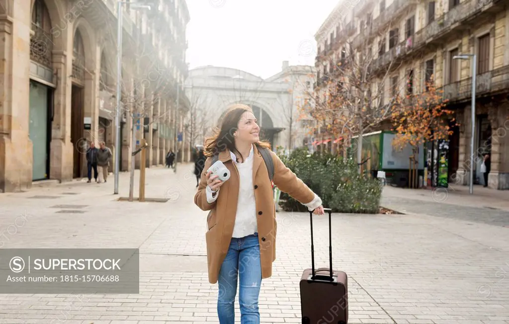 Smiling young woman with suitcase and camera in the city on the go, Barcelona, Spain