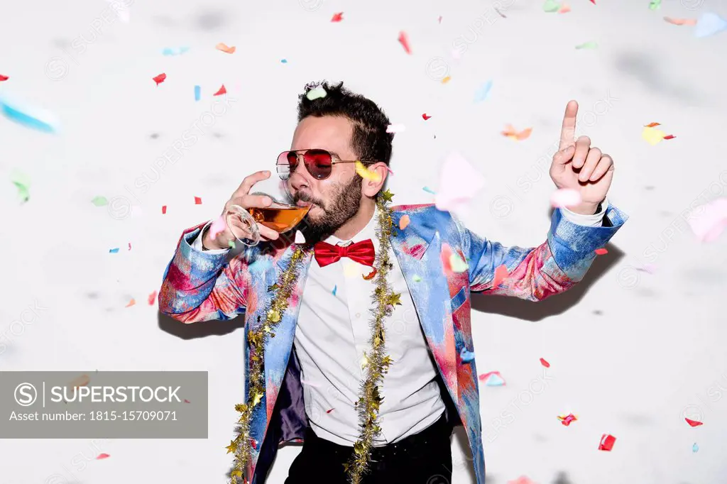 Cool and stylish man wearing a colorful suit and sunglasses celebrating a party with confetti and drinking