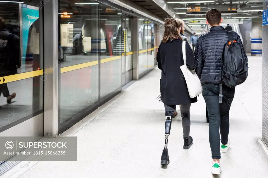 Rear view of young woman with leg prosthesis and man walking at subway station platfom