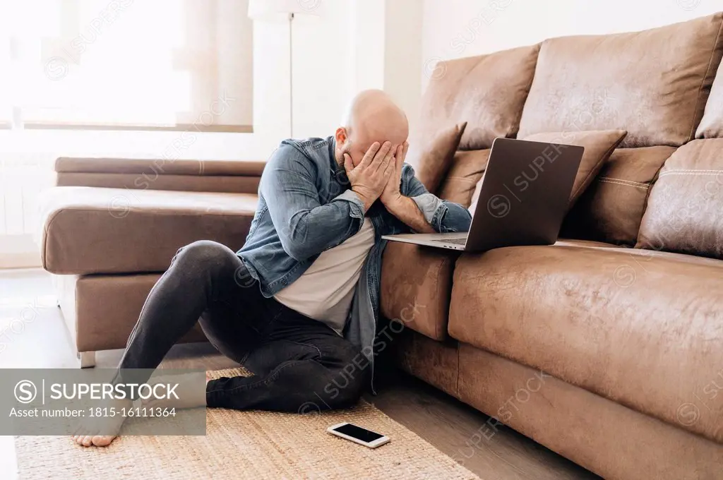 Tired businessman with laptop covering face while sitting in living room at home