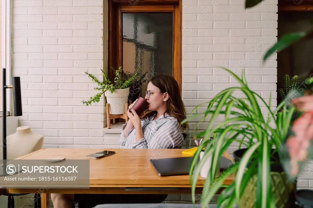 Thoughtful woman drinking coffee while sitting at desk in home office