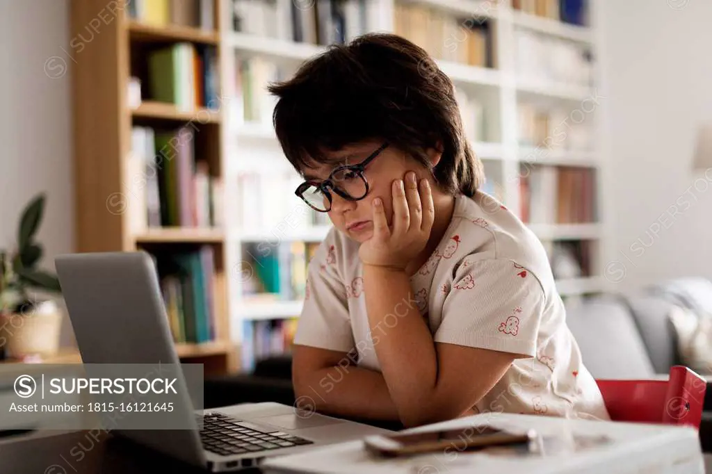 Cute boy with hand on chin looking at laptop while sitting in living room