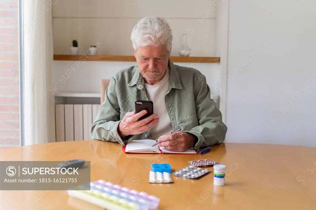 Senior man using smart phone while sitting with diary and medicines on table at home