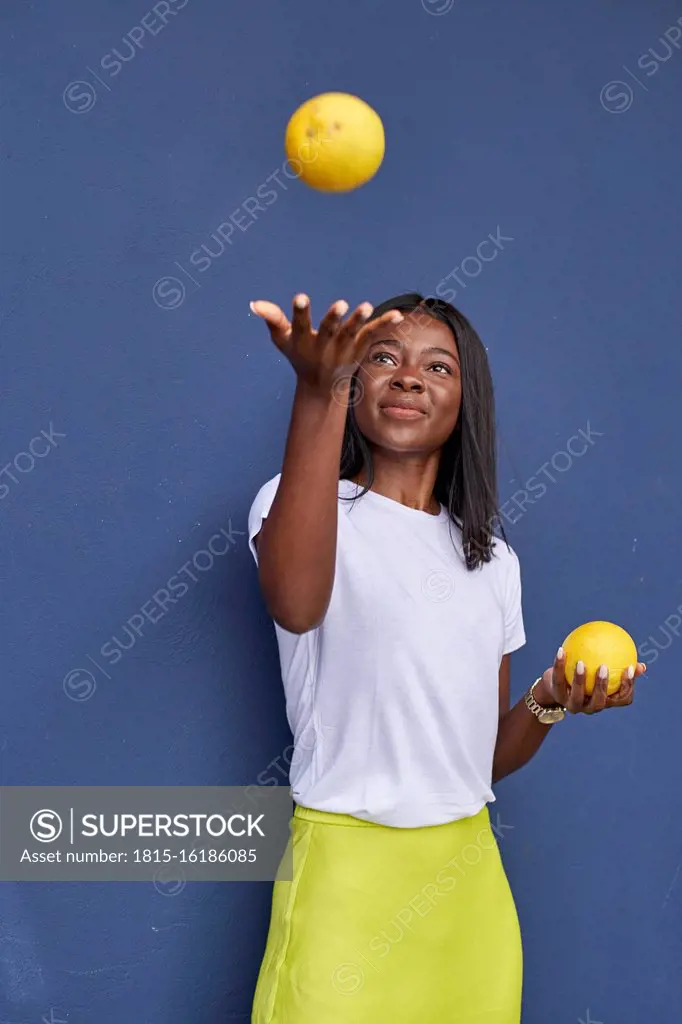 Portrait of happy young woman juggling with two oranges in front of blue background