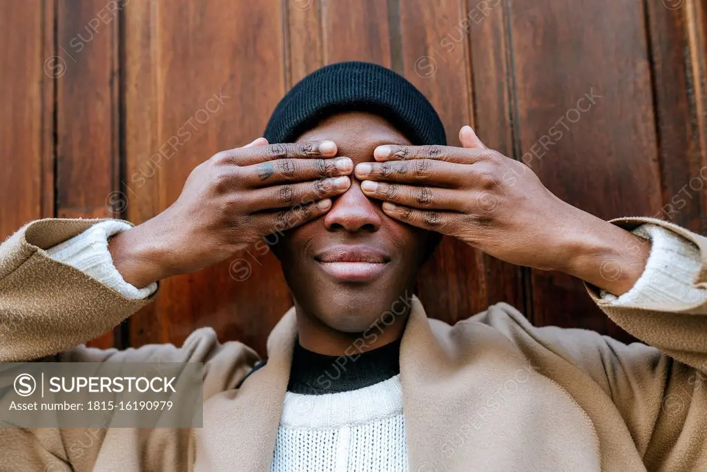 Young woman covering eyes with hands against wooden wall