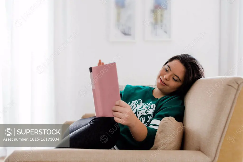 Happy young woman with down syndrome reading book while resting on armchair at home