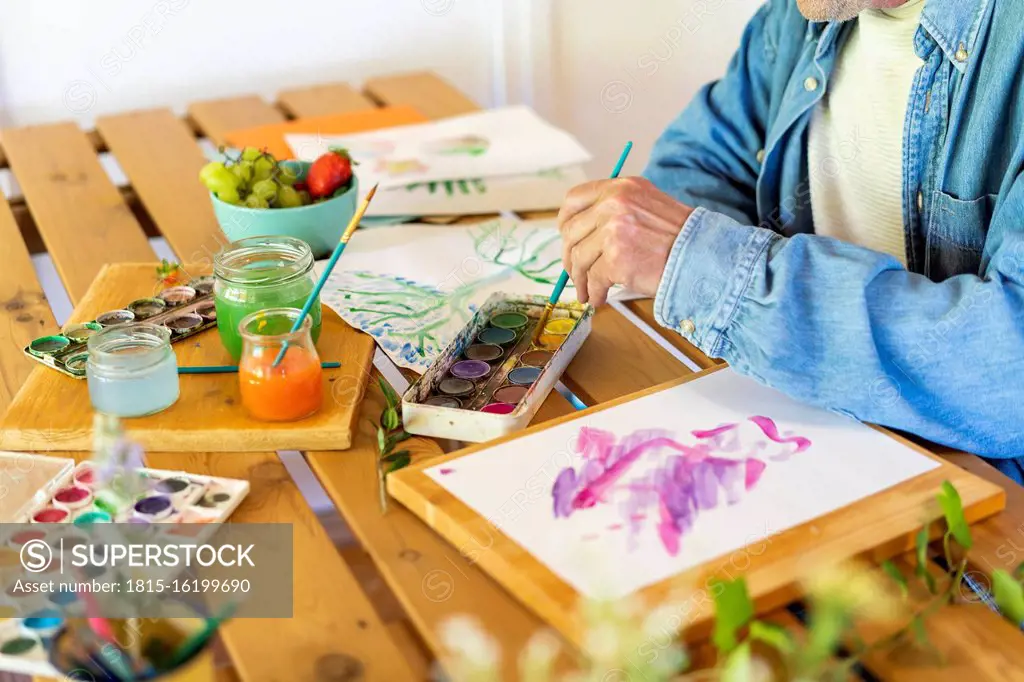 Senior man dipping paintbrush in watercolor paints on table at home