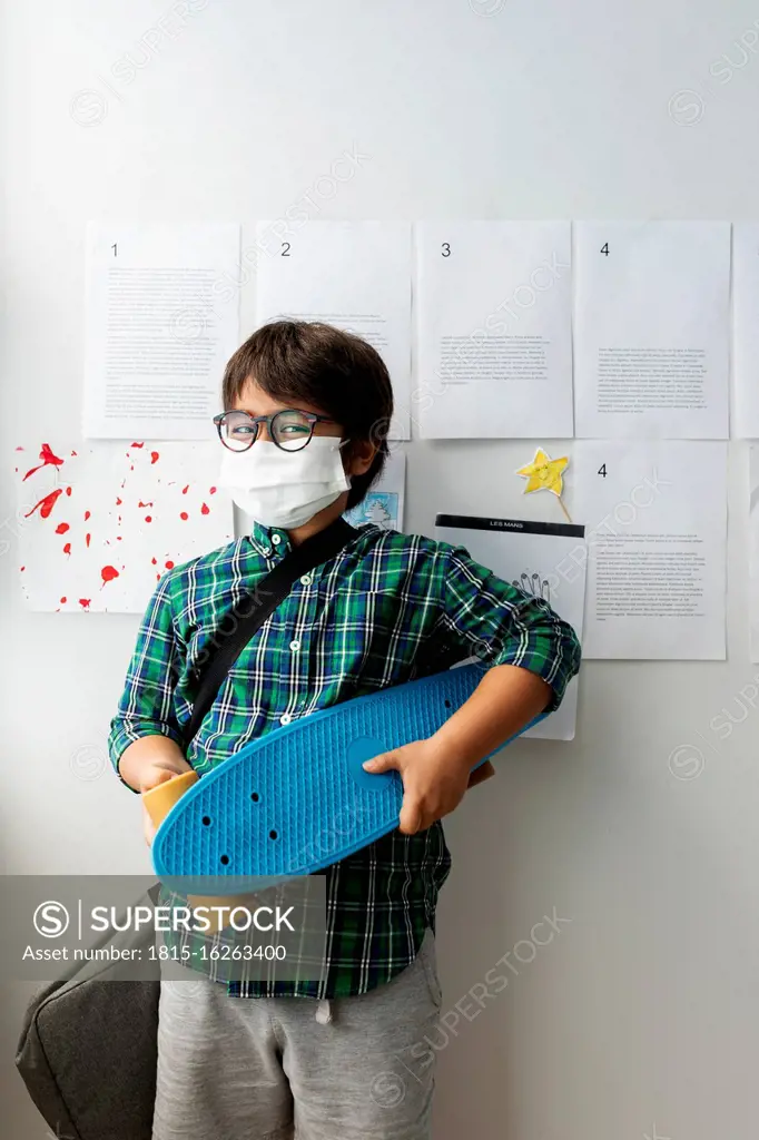 Boy wearing mask holding skateboard while standing against wall in school