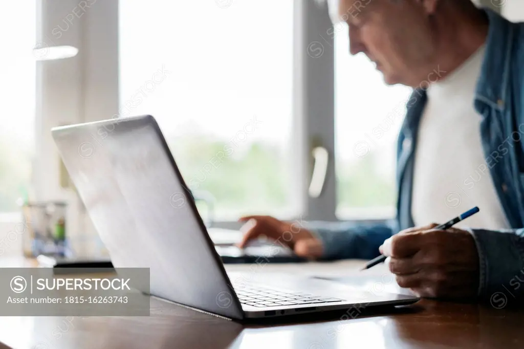 Senior man using calculator and laptop while writing at home