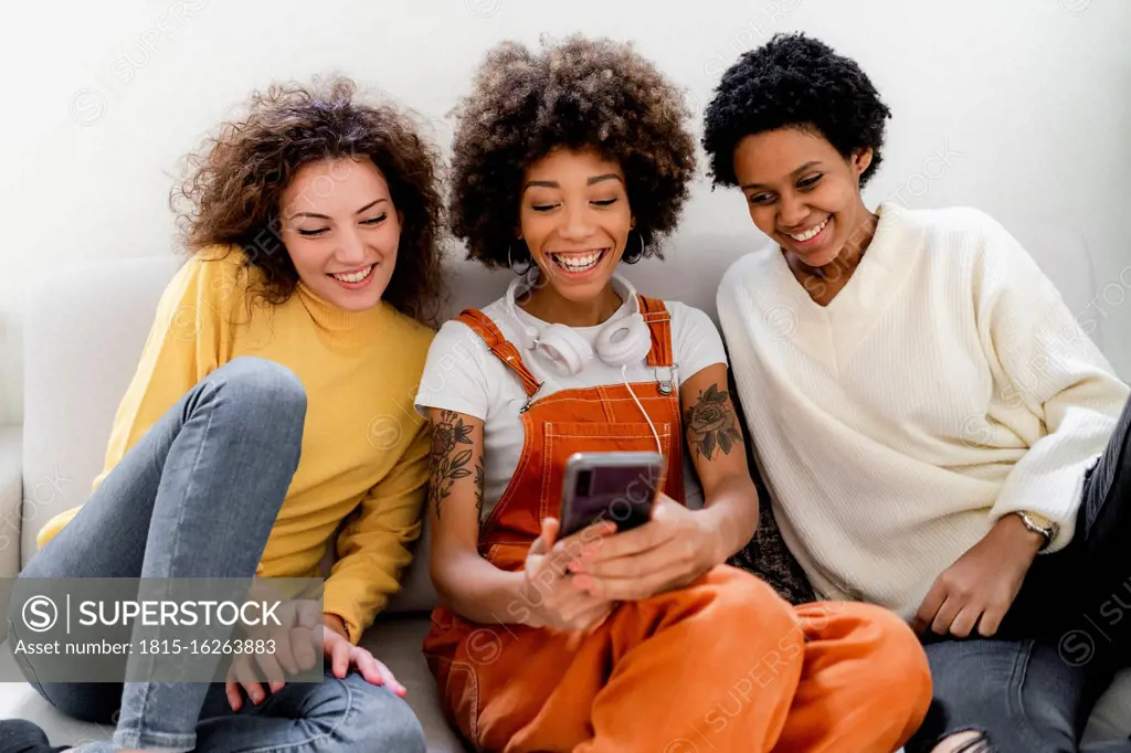 Group picture of three laughing friends sitting on couch taking selfie with smartphone